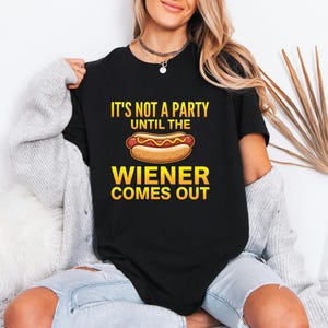 May include: Black t-shirt with the text "IT'S NOT A PARTY UNTIL THE WIENER COMES OUT" in yellow and white. A cartoon hot dog with mustard is centered below the text. The shirt is a casual style.