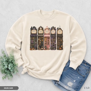May include: A sand-coloured crewneck sweatshirt featuring a graphic print of five storefronts labelled Midnight Potion Bar, Phantom Library, Spellbound Sweets, Moonlit Petals, and Raven Relics, with detailed illustrations of books, potions, and pumpkins.