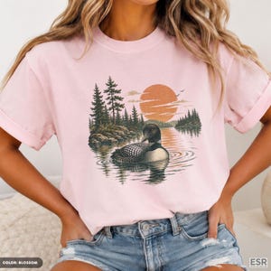 Peut inclure: Un t-shirt rose clair à manches courtes orné d'une illustration représentant un plongeon nageant sur un lac. Le motif comprend un grand soleil orange, une forêt de pins et des rives rocheuses, formant une scène naturelle détaillée.