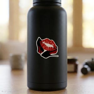 May include: Black water bottle with a sticker featuring a red and white lipstick kiss design. The sticker also includes a black high heel shoe. The background is blurred.