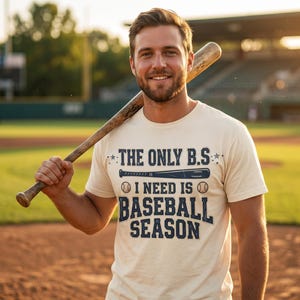 May include: Cream-colored t-shirt with the text "THE ONLY B.S. I NEED IS BASEBALL SEASON" in navy blue. A baseball bat rests on the shoulder. The image is taken at a baseball field.