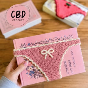 May include: A pink crocheted book cover featuring a small white crocheted bow in the centre and a white crocheted border. The cover is wrapped around a copy of the book Pride and Prejudice by Jane Austen, which has a pink cover with floral art.