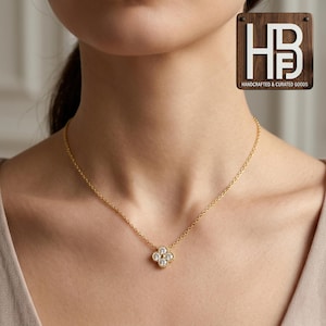 May include: A gold necklace with a four-leaf clover pendant set with white gemstones. The necklace is worn around a person's neck. The image also includes a wooden sign with the text "HANDCRAFTED & CURATED GOODS".