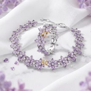 May include: A silver bracelet and ring set with a floral design. The jewellery features purple flower-shaped stones and small gold accents. The set is displayed on a white surface with scattered purple crystals and flowers.