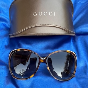 May include: Tortoiseshell-patterned sunglasses with dark-tinted lenses and a matching brown hard-shell case featuring the word GUCCI embossed in gold lettering on the front. The frames have a rounded shape with a smooth, polished finish.