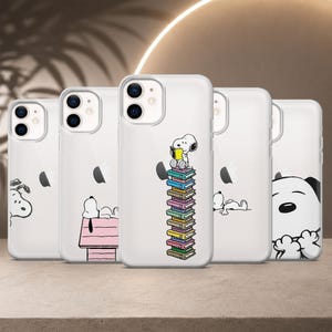 Op de afbeelding: Vijf doorzichtige smartphonehoesjes met diverse Snoopy-illustraties. Eén hoesje toont Snoopy lezend op een stapel boeken, een ander Snoopy op een roze hondenhok, één Snoopy liggend, één Snoopy die om de hoek kijkt en één een close-up van Snoopy.
