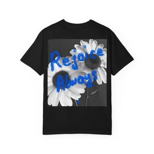 以下が含まれることがあります： 黒いTシャツ。白と黒の2つのヒナギクのグレースケール画像が特徴です。ヒナギクは白い花びらと濃い中心部を持っています。「Rejoice Always」の文字が青色で花の上に書かれています。Tシャツはクルーネックです。
