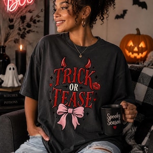 Puede incluir: Una camiseta de manga corta gris carbón con un gráfico rojo que dice "TRICK OR TEASE" junto a cuernos de diablo, un par de labios rojos y un gran lazo rosa claro. La camiseta se combina con unos vaqueros de mezclilla azul claro desgastados.