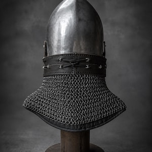 Puede incluir: Un casco de acero pulido con una corona puntiaguda y una superficie lisa. Una banda ancha de cuero negro con tachuelas de metal plateado y un cierre central de cordones rodea la base, unida a una camal de cota de malla gris oscuro.