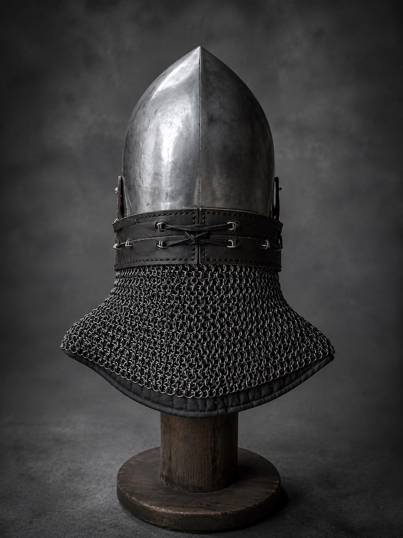 Puede incluir: Un casco de acero pulido con una corona puntiaguda y una superficie lisa. Una banda ancha de cuero negro con tachuelas de metal plateado y un cierre central de cordones rodea la base, unida a una camal de cota de malla gris oscuro.
