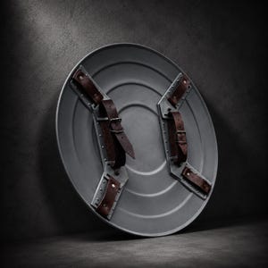 May include: A circular gray metal shield featuring a smooth, concentric ring design. The interior side is equipped with two metallic mounting brackets, each secured with rivets and fitted with adjustable dark brown leather straps and buckles.