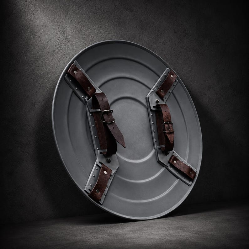 May include: A circular gray metal shield featuring a smooth, concentric ring design. The interior side is equipped with two metallic mounting brackets, each secured with rivets and fitted with adjustable dark brown leather straps and buckles.