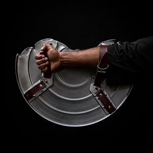 K&ouml;nnte beinhalten: Ein kreisf&ouml;rmiger Metallschild mit glattem, konzentrischem Ringdesign und silberner Oberfl&auml;che. Der Schild verf&uuml;gt &uuml;ber zwei dunkelbraune Lederriemen mit Metallschnallen und Nieten, die zur Befestigung am Unterarm und f&uuml;r einen Griff dienen.