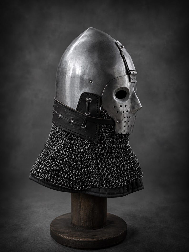 Puede incluir: Un casco de acero pulido con una m&aacute;scara facial articulada que presenta aberturas para los ojos y orificios de ventilaci&oacute;n. El casco incluye un protector de cuello de cuero negro asegurado con remaches met&aacute;licos y una cota de malla.