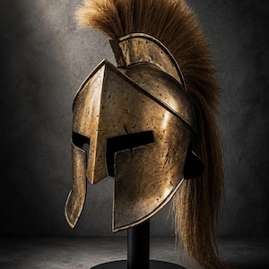 Puede incluir: Un casco de estilo espartano de color bronce con un acabado desgastado y envejecido, que presenta una cresta alta y prominente hecha de pelo largo, grueso y de color marrón claro que se extiende desde la parte superior hasta la parte posterior.
