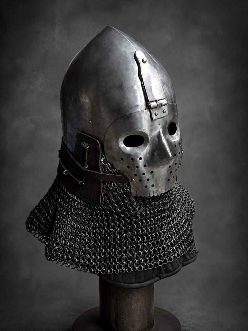 Puede incluir: Un casco de acero pulido con una c&uacute;pula redondeada, una placa facial articulada con dos aberturas para los ojos y una zona perforada para la nariz y la boca. Una correa de cuero oscuro asegura la estructura met&aacute;lica a una cota de malla.