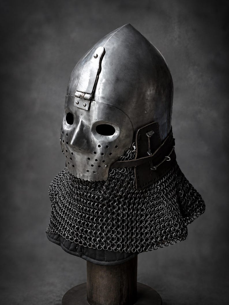 Puede incluir: Un casco de acero pulido con una placa facial articulada que presenta aberturas para los ojos y orificios de ventilaci&oacute;n. El casco incluye una correa de cuero oscuro y una cota de malla protectora que cubre el cuello y la parte inferior.