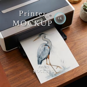 Può includere: Una stampante da scrivania nera e argento con un foglio di carta che mostra un'illustrazione ad acquerello di un airone cenerino. Il testo "Printer MOCKUP", "4K", "Mockzi" e "MOCKUPS" è visibile sull'immagine e sulla superficie della stampante.