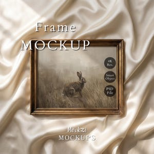 May include: A framed art print featuring a brown rabbit in a field, set within a gold-colored ornate frame. The words "Frame MOCKUP" are at the top, with "Mockzi MOCKUPS" at the bottom. The image is on a cream-colored satin fabric.