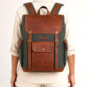 May include: A dark green canvas backpack with brown leather accents, featuring a top flap, two vertical buckled straps, a central front pocket with a snap closure, two side pockets, and a small rectangular label with the text SAFARIEST.