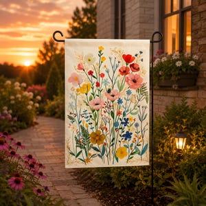 Può includere: Una bandiera da giardino con un vivace design floreale, con fiori rosa, rossi, gialli e blu. La bandiera è montata su un supporto in metallo nero in un giardino, con un tramonto sullo sfondo. La bandiera ha uno sfondo color crema.