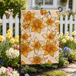 Könnte beinhalten: Eine rechteckige Gartenflagge mit einem sich wiederholenden Muster aus gelben und orangen Narzissen auf cremefarbenem Grund. Das Design enthält Illustrationen von Ketten und Zahnrädern sowie Text in den Blütenmitten mit der Aufschrift Demand Justice und Resist the Hate.