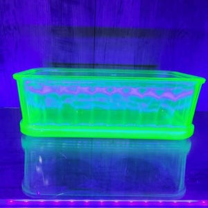 May include: A rectangular, glowing green glass dish with a ribbed design. The dish has a clear, reflective base, and the interior shows a wavy, pinkish pattern. The background is a dark blue.