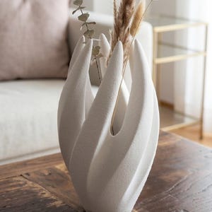 Op de afbeelding: Een witte, sculpturale vaas met een uniek, organisch ontwerp. De vaas bevat gedroogde decoratieve grassen en bladeren. De vaas staat op een houten tafel. Een kaart op de tafel luidt "THE ECLIPSE VASE - Artisanal 3D Sculpture."