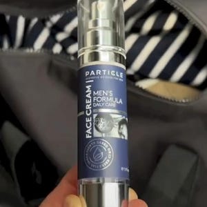 May include: A silver and blue Particle face cream bottle. The label reads "FACE CREAM" and "MEN'S FORMULA DAILY CARE". The bottle has a clear cap and a pump dispenser. The background is a dark grey bag.