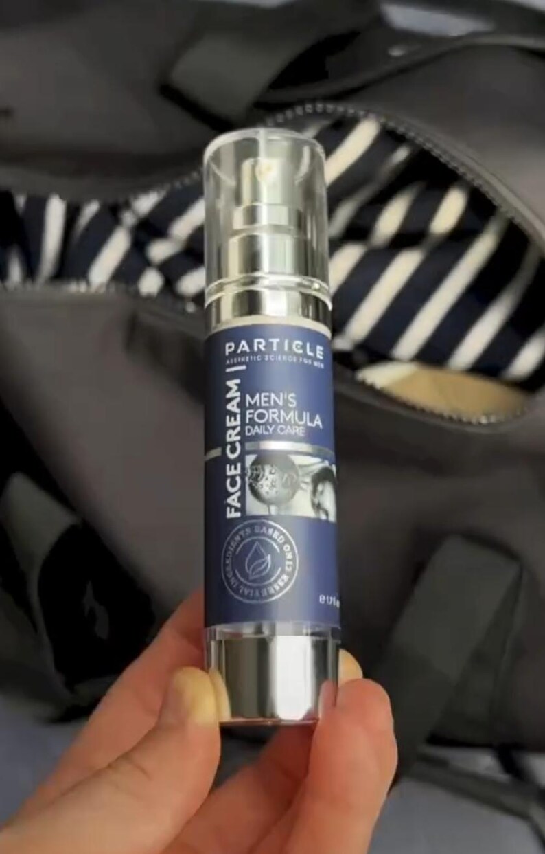 May include: A silver and blue Particle face cream bottle. The label reads "FACE CREAM" and "MEN'S FORMULA DAILY CARE". The bottle has a clear cap and a pump dispenser. The background is a dark grey bag.