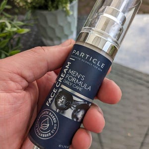 May include: A dark blue and silver face cream bottle with the text "PARTICLE" and "MEN'S FORMULA DAILY CARE". The bottle has a clear plastic cap and is held in a hand. The bottle contains 50 ml.