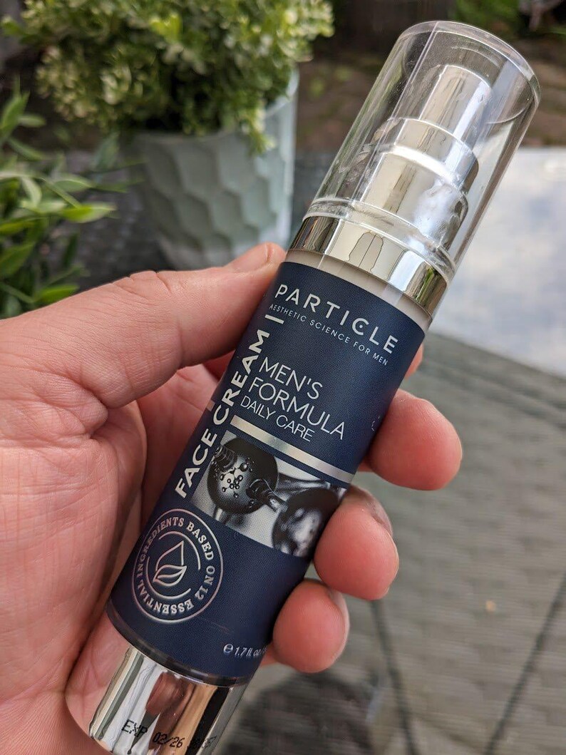 May include: A dark blue and silver face cream bottle with the text "PARTICLE" and "MEN'S FORMULA DAILY CARE". The bottle has a clear plastic cap and is held in a hand. The bottle contains 50 ml.