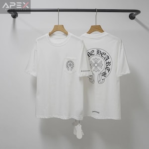 May include: Two white t-shirts hanging on a rack. One shirt has a pocket with a black design, and the other has a large black graphic on the back. The shirts are made of a soft material and have short sleeves.