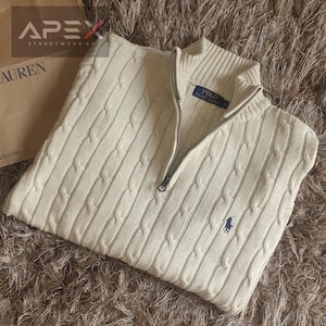May include: Cream cable-knit sweater with a quarter-zip. The sweater has a ribbed collar and cuffs, and a small navy blue polo player logo. The garment is folded on a textured surface.