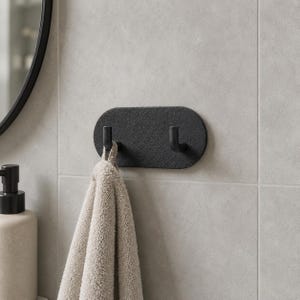 May include: A black, oval-shaped wall hook with two L-shaped hooks. A beige towel hangs from one hook. The hook is mounted on a light gray tiled wall. A black mirror and soap dispenser are visible in the background.