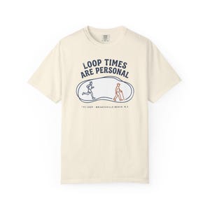 Puede incluir: Una camiseta de manga corta color crema con un gráfico azul oscuro y el texto LOOP TIMES ARE PERSONAL. El diseño incluye un bucle de líneas con un corredor y un caminante, y el texto THE LOOP - WRIGHTSVILLE BEACH, N.C. en la parte inferior.