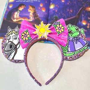Peut inclure: Un serre-tête rose à paillettes avec un grand nœud rose orné d'un soleil jaune et de fleurs. Le serre-tête comporte des images de dessins animés d'un cheval blanc, d'un caméléon vert et de fleurs. L'arrière-plan montre une illustration de livre avec un bateau et des lanternes.