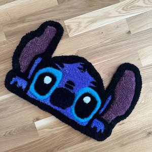 Könnte beinhalten: Handgefertigter Teppich in Form der Disney-Figur Stitch. Der Teppich hat einen blauen Körper, eine schwarze Umrandung und lila Ohren. Die Augen sind groß und weiß mit schwarzen Pupillen. Der Teppich liegt auf einem Holzboden.