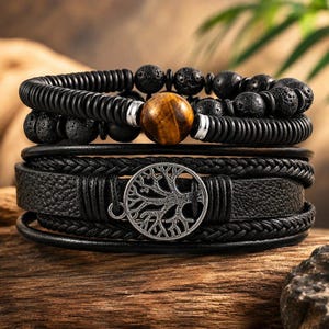 May include: A multi-layered bracelet set featuring a strand of black textured beads and a single brown tiger eye bead, paired with a black leather strap bracelet that includes a silver-toned circular charm depicting a detailed tree design.