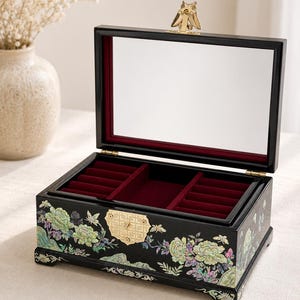 May include: Black jewelry box with an open lid, featuring a mirror and red velvet lining. The box is adorned with floral and butterfly designs in shades of green, white, and iridescent accents. A gold-colored owl-shaped clasp secures the lid.