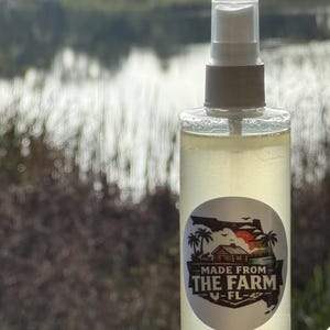 May include: A transparent spray bottle with a white and brown cap, containing a yellow liquid. The bottle features a round sticker with the text "MADE FROM THE FARM - FL" and a graphic of a house, palm trees, and the state of Florida.