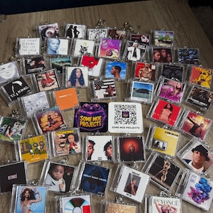 Puede incluir: Una colección de llaveros acrílicos transparentes con portadas de álbumes de varios artistas musicales. Los llaveros muestran portadas de álbumes con diversos colores y diseños, incluyendo texto e imágenes. Algunos llaveros tienen anillos de metal plateado.