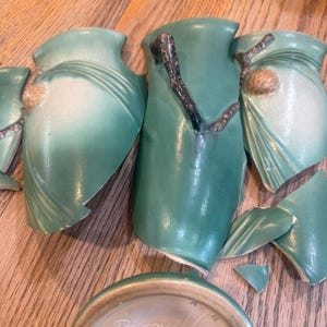 May include: Broken pieces of a green ceramic vase with raised brown branch and pinecone details. The base is visible, featuring the stamped text Roseville 843-8. The ceramic has a matte finish with varying shades of green and brown accents.