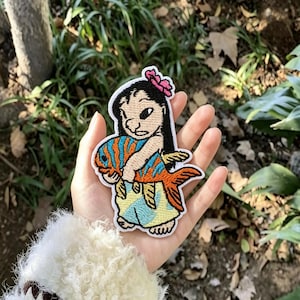 May include: An embroidered patch featuring a cartoon character with black hair and a pink flower, wearing a light blue skirt, holding a large orange and blue striped fish. The patch has a white border and detailed stitching throughout the design.