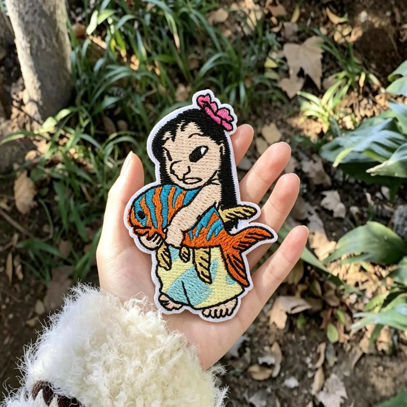 May include: An embroidered patch featuring a cartoon character with black hair and a pink flower, wearing a light blue skirt, holding a large orange and blue striped fish. The patch has a white border and detailed stitching throughout the design.