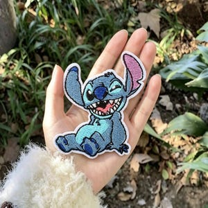 May include: An embroidered patch featuring the character Stitch from Lilo & Stitch. The patch is light blue with a darker blue nose, inner ears, and paws. It shows the character sitting with a wide, toothy grin, one ear upright, and one ear folded.