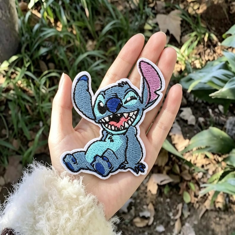 May include: An embroidered patch featuring the character Stitch from Lilo & Stitch. The patch is light blue with a darker blue nose, inner ears, and paws. It shows the character sitting with a wide, toothy grin, one ear upright, and one ear folded.