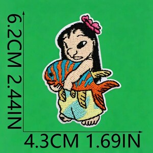 May include: An embroidered patch featuring a character with black hair and a pink flower, holding an orange fish with blue stripes. The character wears a light blue skirt. The patch measures 6.2 cm by 4.3 cm, or 2.44 inches by 1.69 inches.