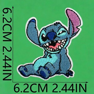 May include: An embroidered patch featuring a blue alien character with large ears, a wide grin, and sharp teeth. The patch measures 6.2 cm or 2.44 inches in height and width, as indicated by the black measurement lines printed on the image.