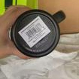 Peut inclure: Un mug noir avec une anse, vu par le dessous. Une &eacute;tiquette rectangulaire blanche est appos&eacute;e sur la base, pr&eacute;sentant un code-barres, le texte 'MADE IN CHINA' et d'autres petits d&eacute;tails imprim&eacute;s. Le mug a une base circulaire et une finition sombre.